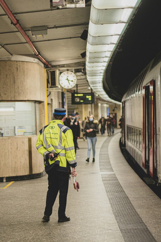 Bahnhofsplattform mit einem Bahnmitarbeiter in einer leuchtend gelben Sicherheitsweste, der mit dem Rücken zur Kamera steht. Ein Zug ist auf der rechten Seite zu sehen, und andere Fahrgäste gehen im Hintergrund in der Nähe einer großen Wanduhr. Dies ist einer der Mitarbeiter, die am Management von Bahnunfällen beteiligt sind.