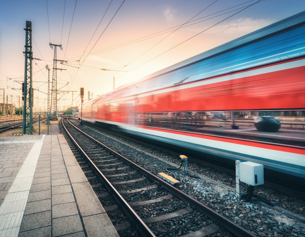 A high-speed red and silver train blurs past a railway station platform at sunset, capturing the motion and speed of travel, with power lines and tracks extending into the distance.